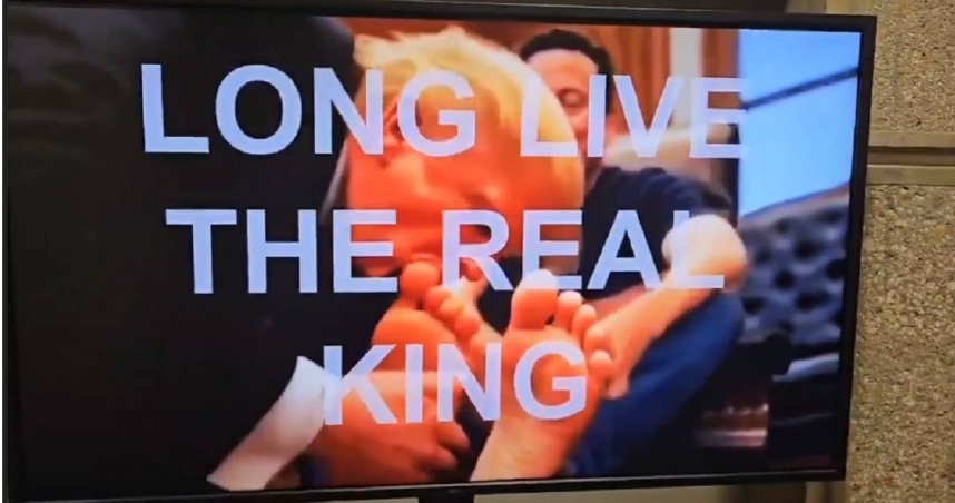 24日，美國「住房與城市發展部」大樓內的電視突然播放了一段AI短片，片中川普跪地親吻馬斯克的腳趾，螢幕上還打上字幕「真王萬歲」（LONG LIVE THE REAL KING），諷刺意味十分濃厚。（圖／翻攝X）